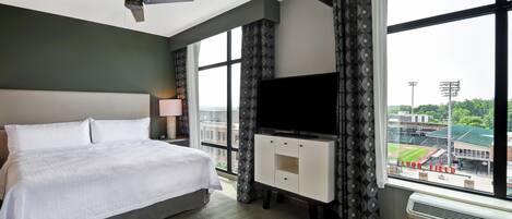 A hotel room with a bed, a flat-screen TV, a ceiling fan, and a large window offering a view of a stadium.