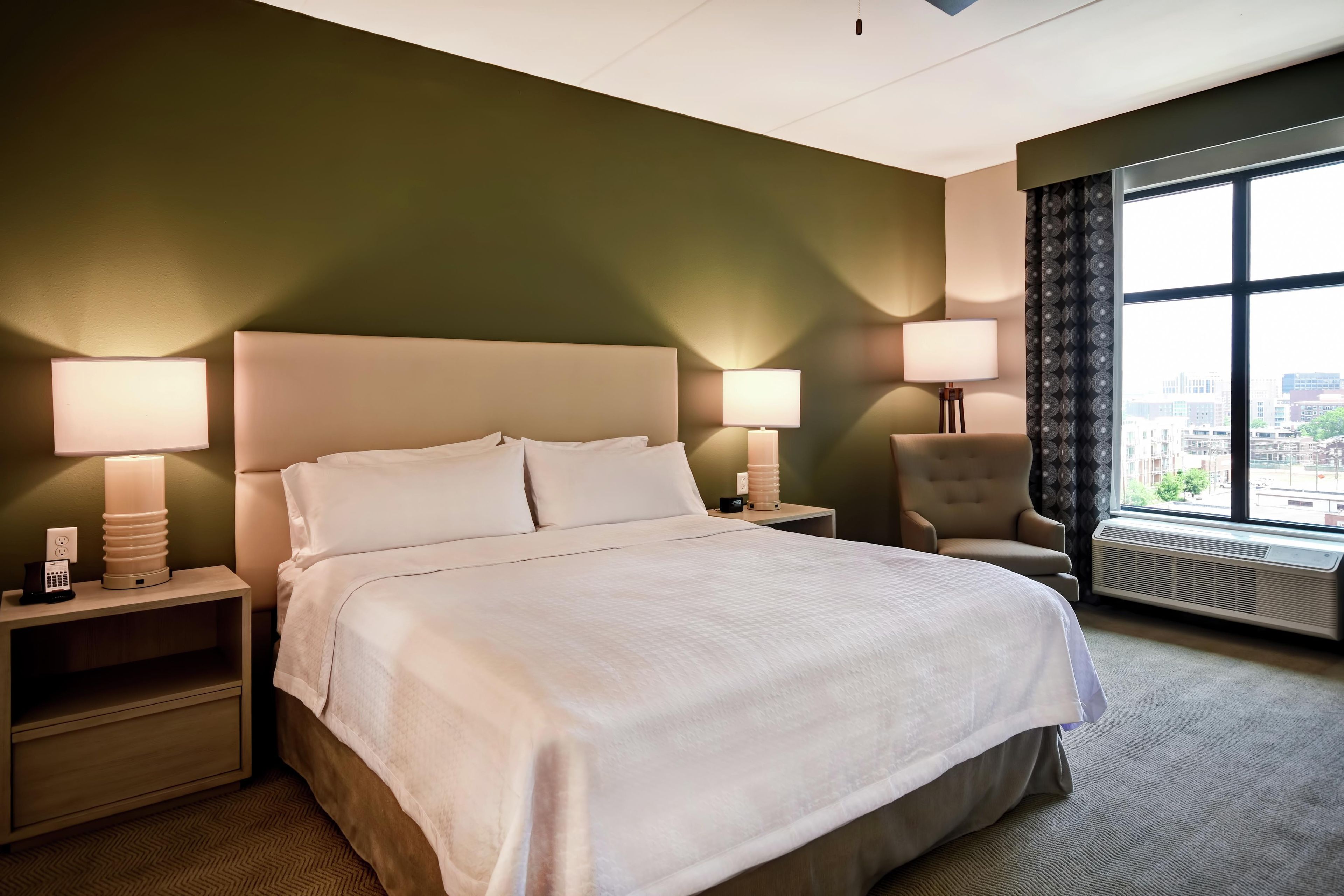 A hotel room with a large bed, two bedside lamps, a chair, a window with a view, and a thermostat.