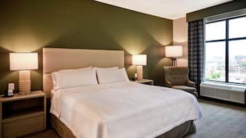 A hotel room with a large bed, two bedside lamps, a chair, a window with a view, and a thermostat.