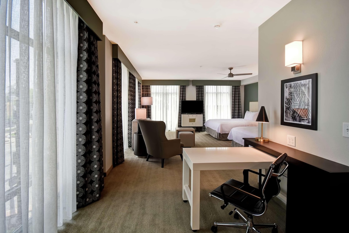 Ein Hotelzimmer mit Schreibtisch, Stuhl, Bett, Fernseher und einem großen Fenster mit Vorhängen.