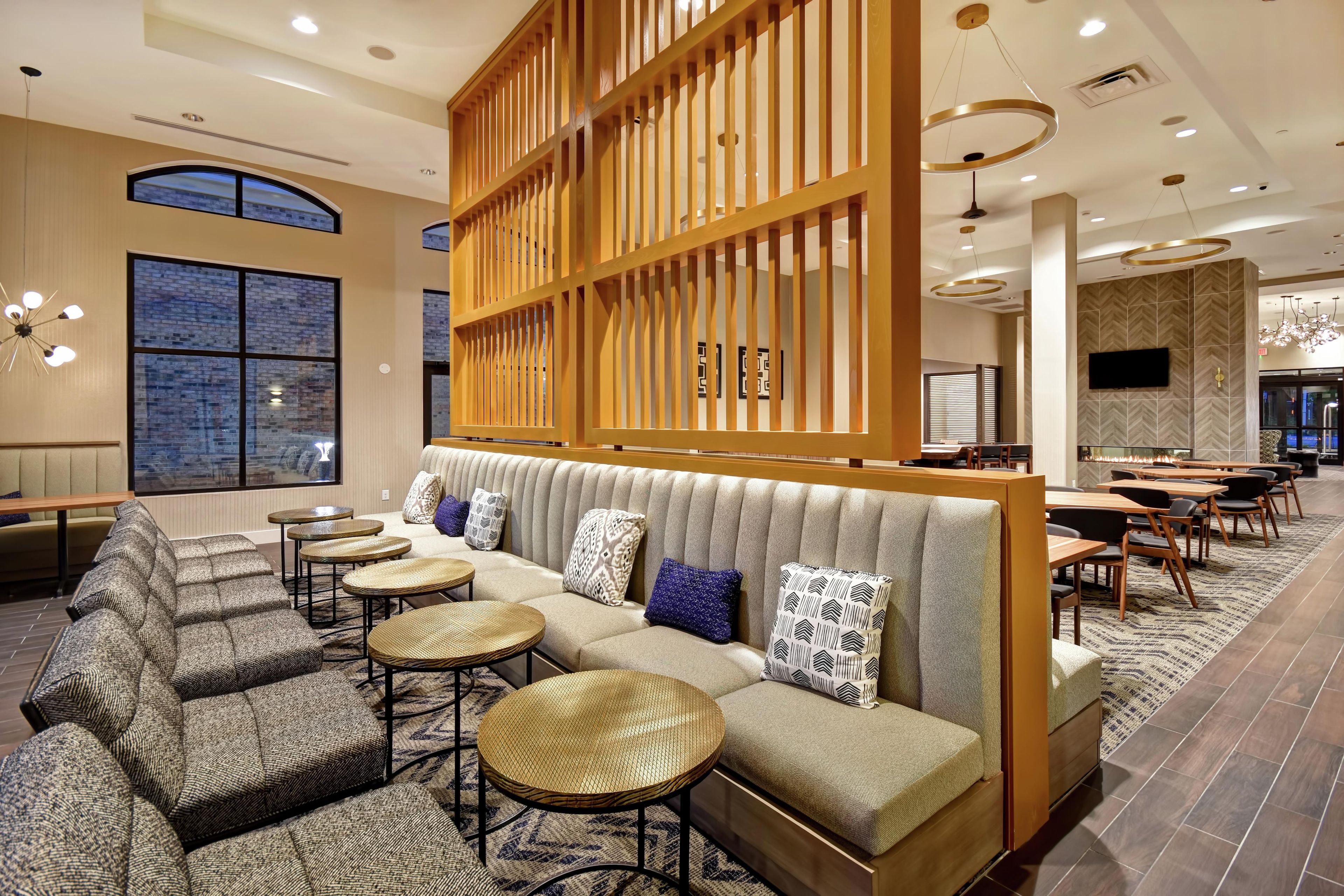 A modern hotel lobby with a seating area featuring a sofa and round tables, large windows, and contemporary lighting fixtures.