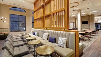 A modern hotel lobby with a seating area featuring a sofa and round tables, large windows, and contemporary lighting fixtures.