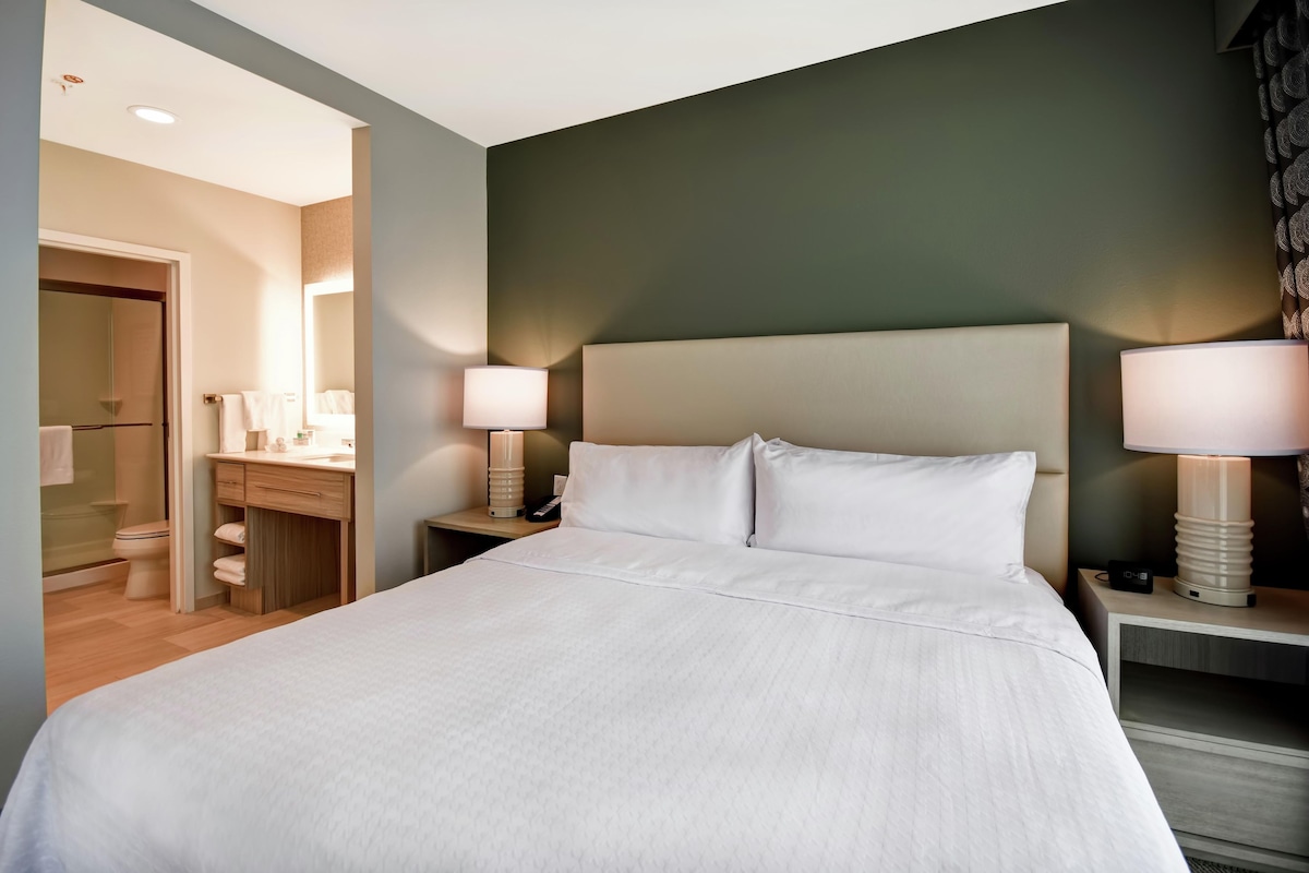 Een hotelkamer met een groot bed, twee nachtkastjes met lampen, een bijzettafeltje met telefoon, en een badkamer zichtbaar door een geopende deur.