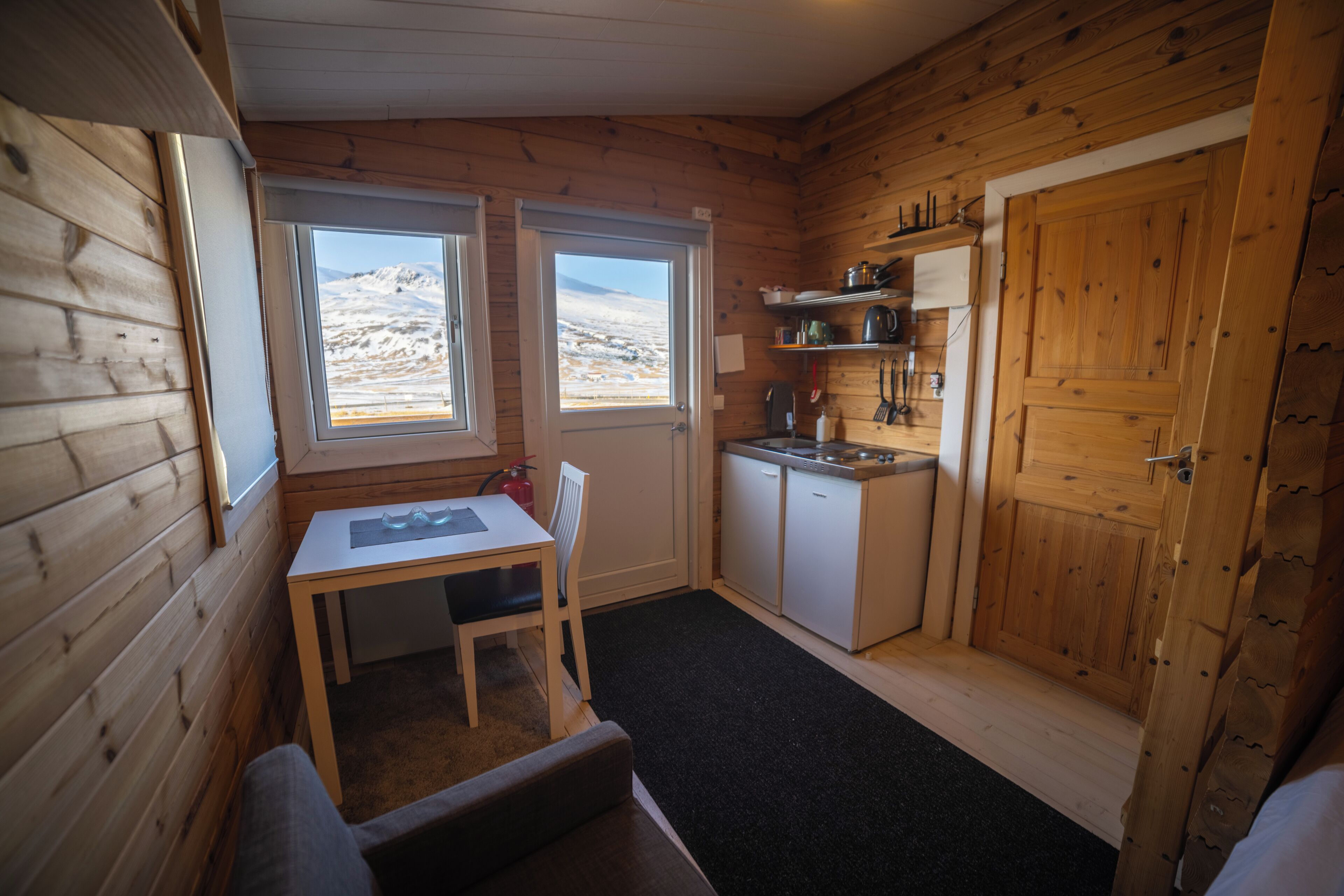 A compact cabin with a wooden interior, a small kitchenette, a dining area, and a view of a snowy landscape through the windows.