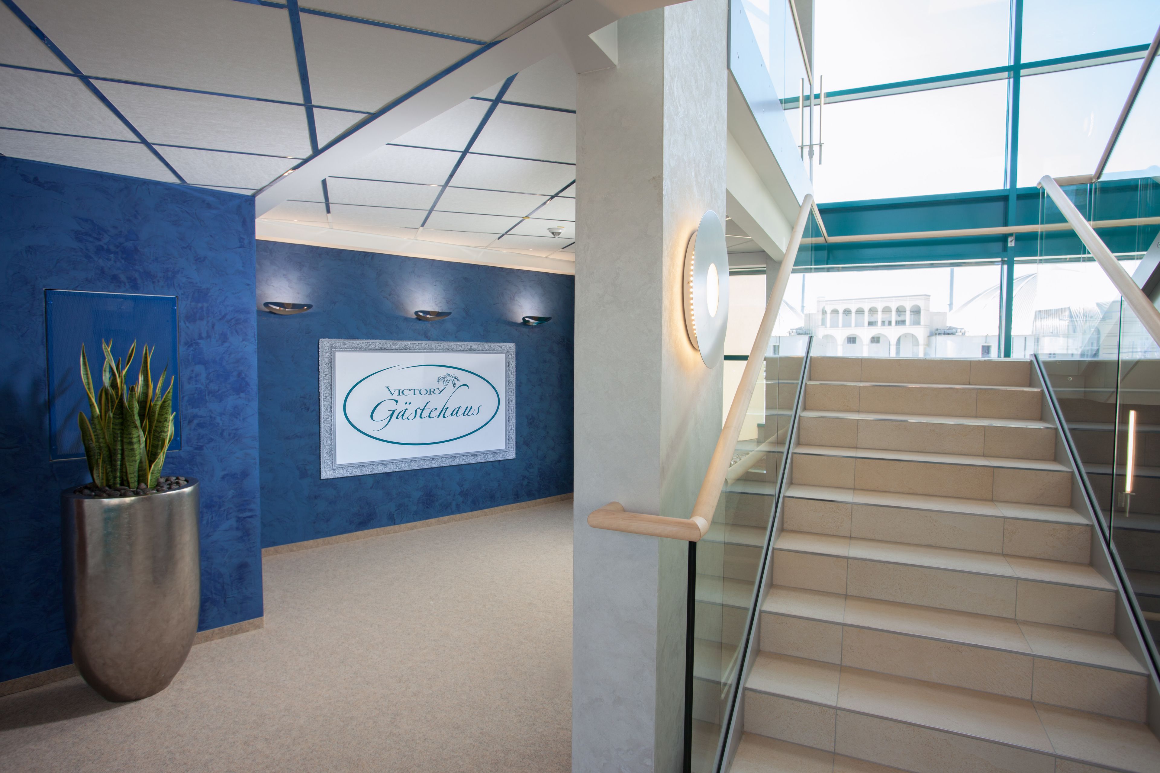 Een modern interieur met een blauwe accentmuur, een lijst met de tekst 'Victory Gasthaus', een grote potplant en een trap met glazen balustrades.