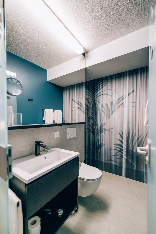 A modern bathroom with a white sink, black faucet, and a toilet. There is a mirror above the sink and a towel rack with two white towels. The walls are painted in a teal color, and there is a curtain with a leaf pattern.