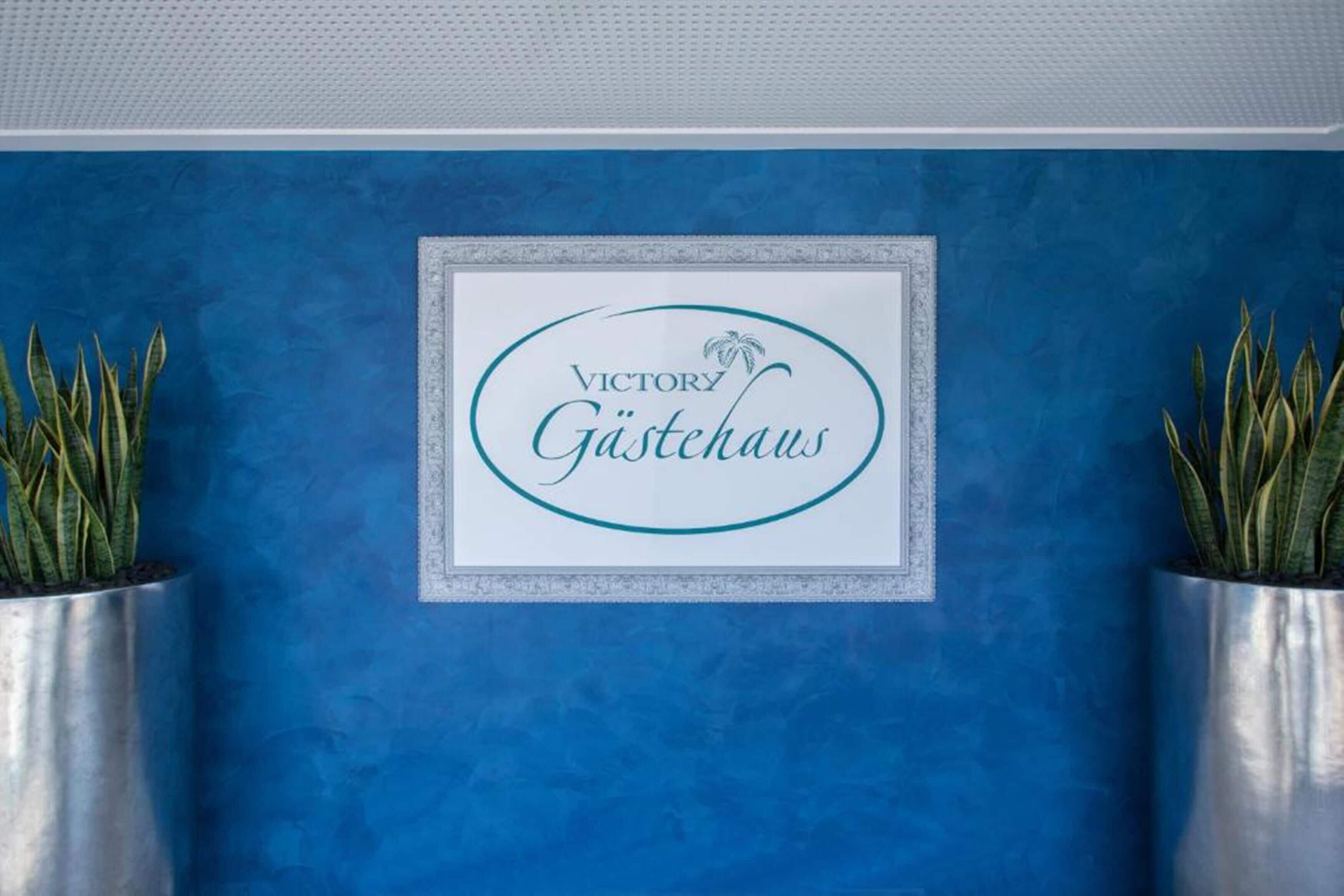 A framed sign with "VICTORY Gästhaus" text on a blue wall, flanked by two potted plants.