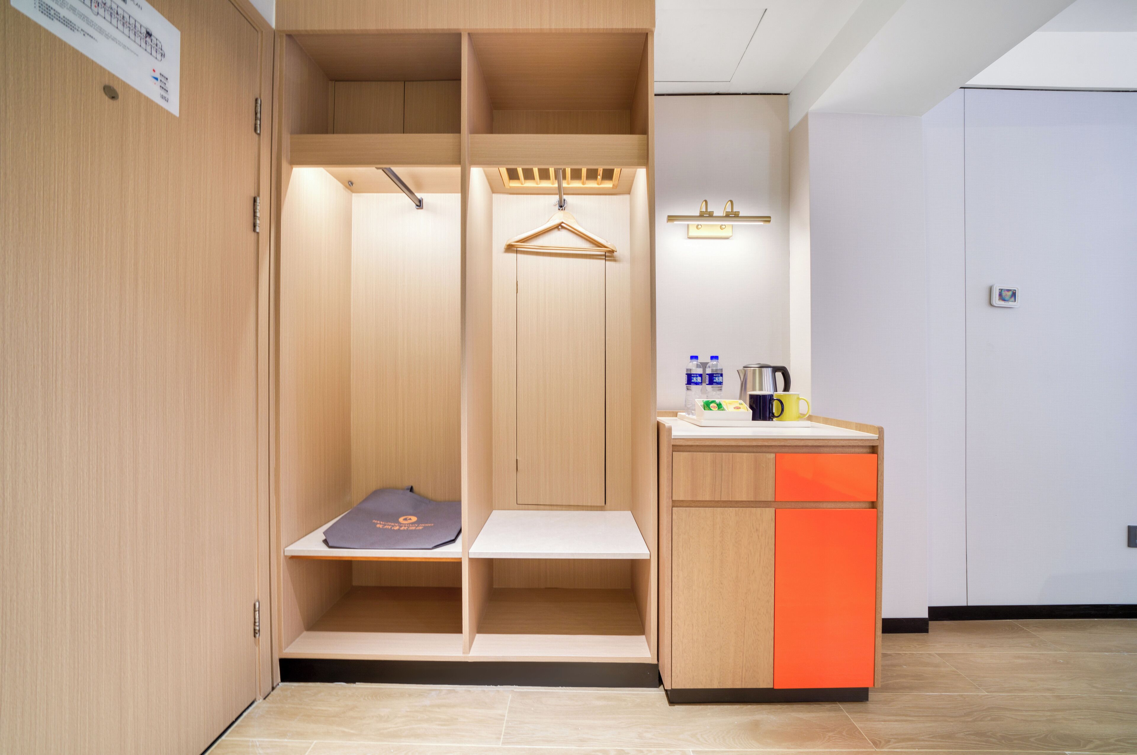 A hotel room with a wooden wardrobe, a small bench, a hanging rack, and a kitchenette with a sink and countertop.