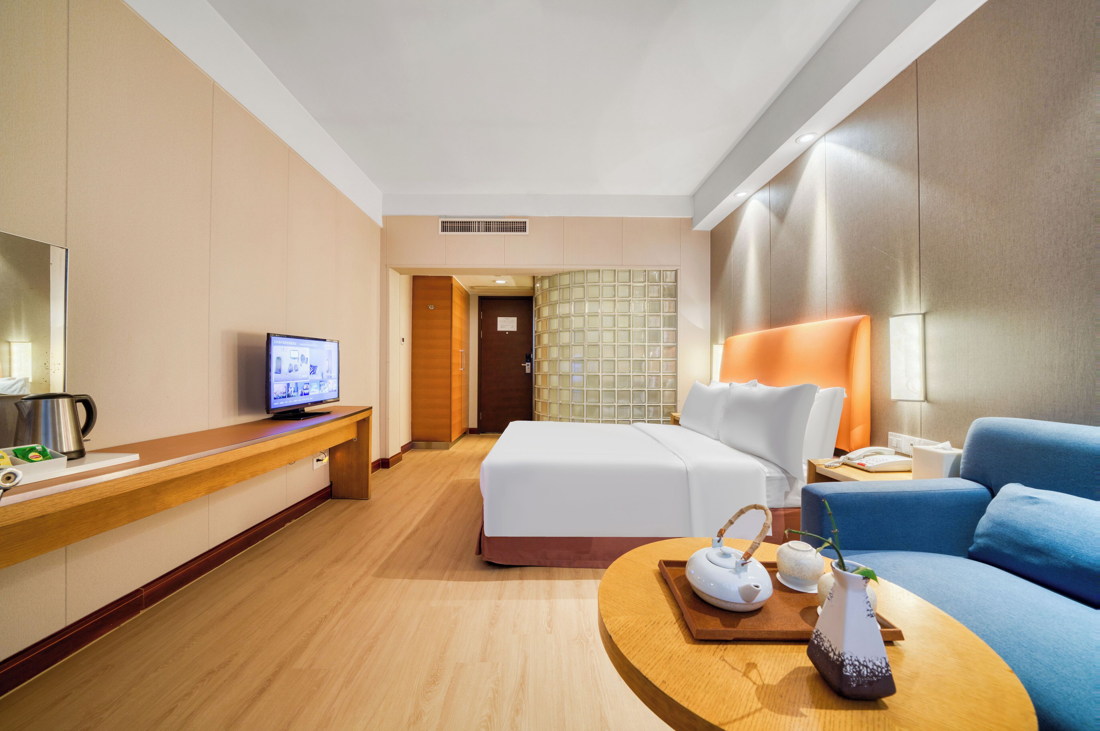 A hotel room with a bed, a desk with a computer, a coffee maker, a lamp, and a vase with a plant.