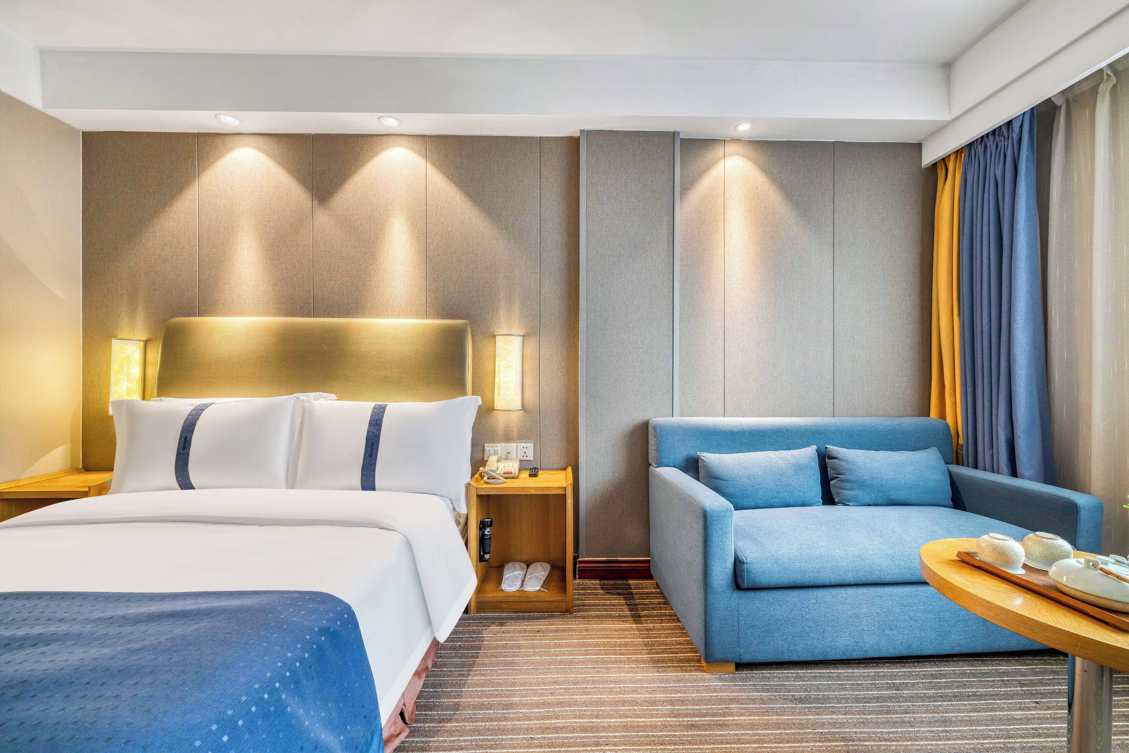 A hotel room with a bed, a sofa, a small table with a tray, and a nightstand with a lamp.
