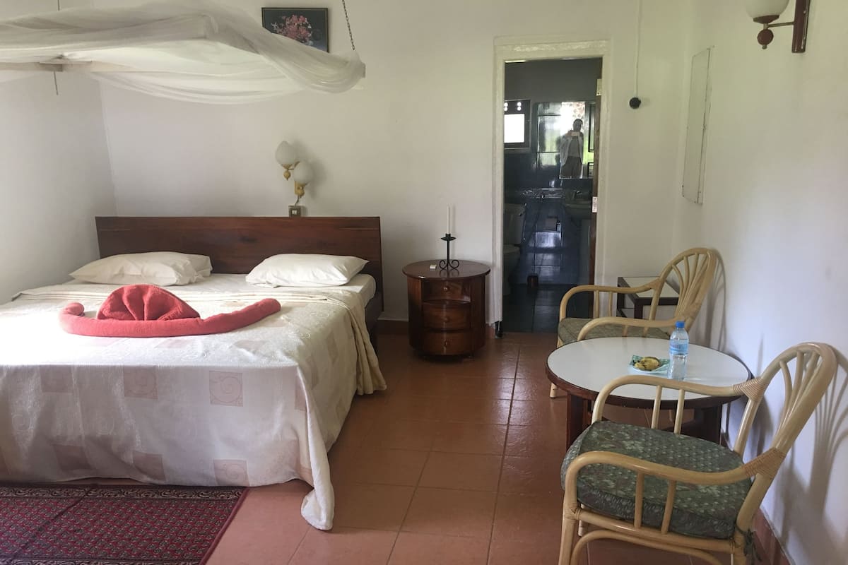 Quarto com cama, uma pequena mesa com cadeiras, mesa de cabeceira, abajur e porta que dá para outro cômodo.