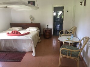 A room with a bed, a small table with chairs, a bedside table, a lamp, and a door leading to another room. - Momella Wildlife Lodge (Arusha)