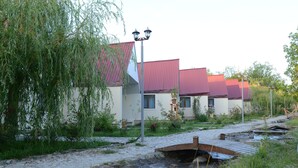 Una hilera de edificios de una sola planta con tejados rojos, un camino adoquinado y un pequeño arroyo.