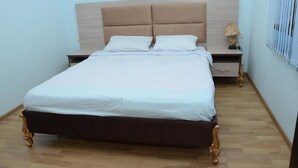 Una cama bien hecha con estructura y cabecera de madera, una mesita de noche con una lámpara y una ventana con persianas.