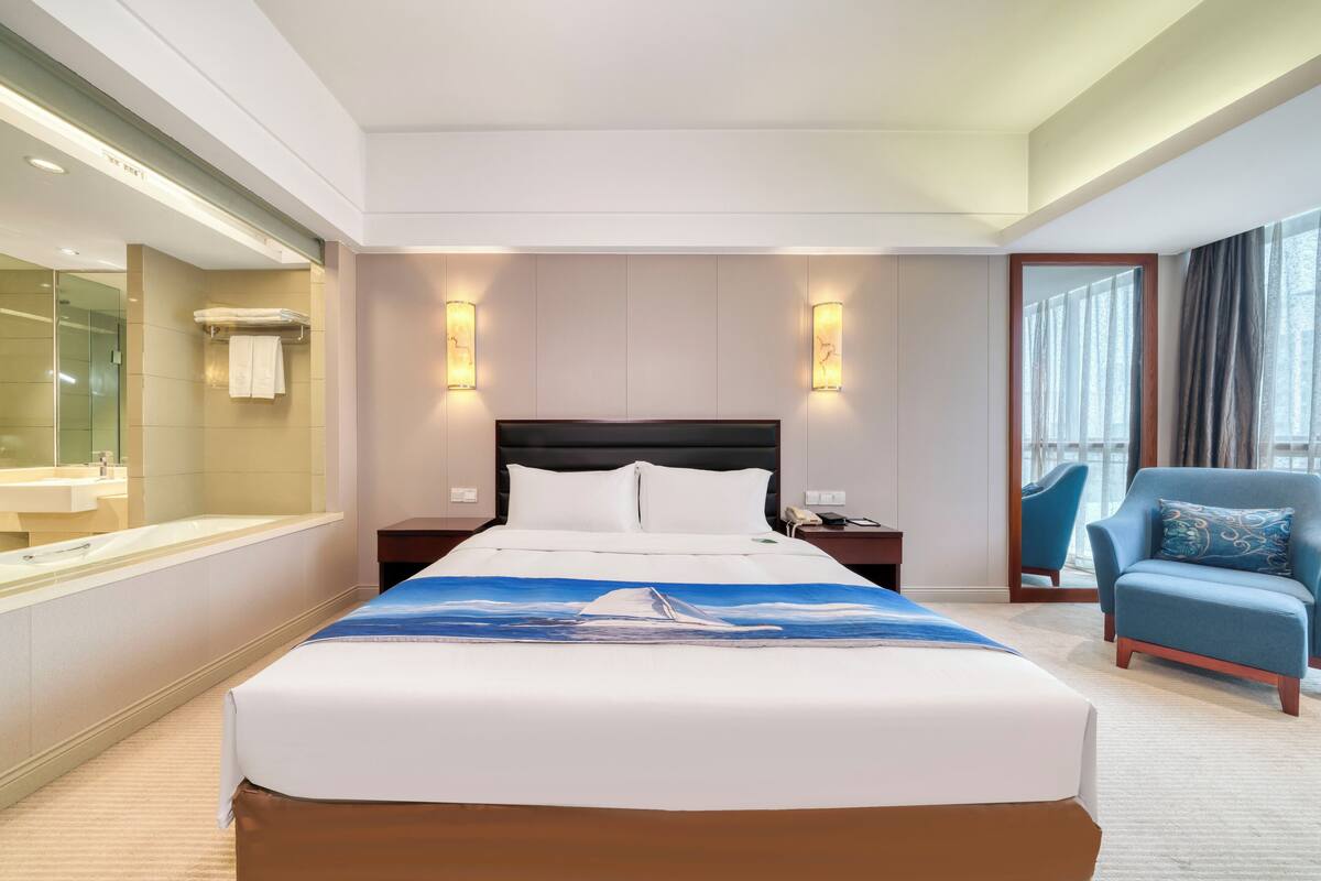 Ein Hotelzimmer mit einem großen Bett, einem Nachttisch, einem Sessel, einem Badezimmer mit einer Badewanne und einem großen Fenster.