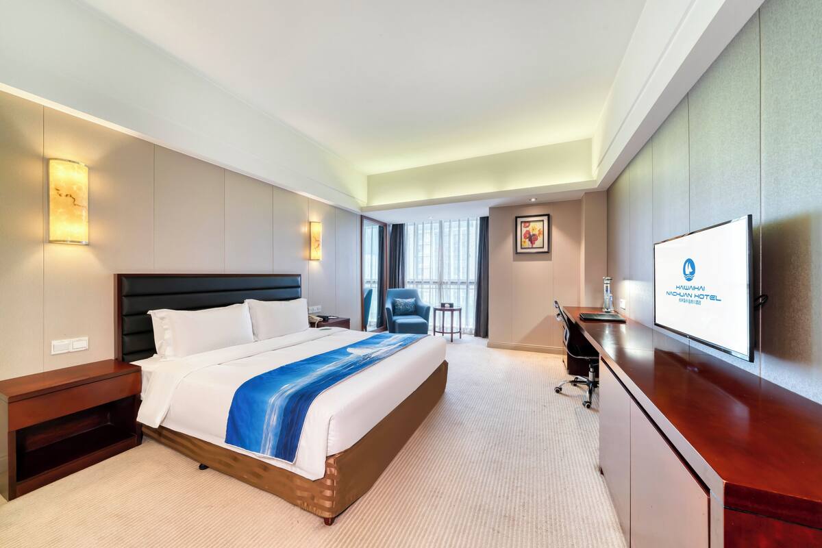 Ein Hotelzimmer mit einem großen Bett, einem Schreibtisch, einem Sessel, einem Fernseher und einem Fenster mit Vorhängen.
