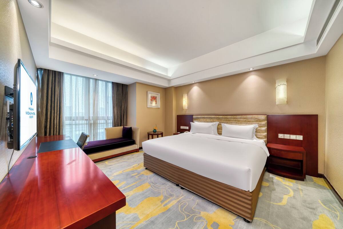 Ein Hotelzimmer mit einem großen Bett, einem Schreibtisch, einem Fernseher und einem Fenster mit Vorhängen.