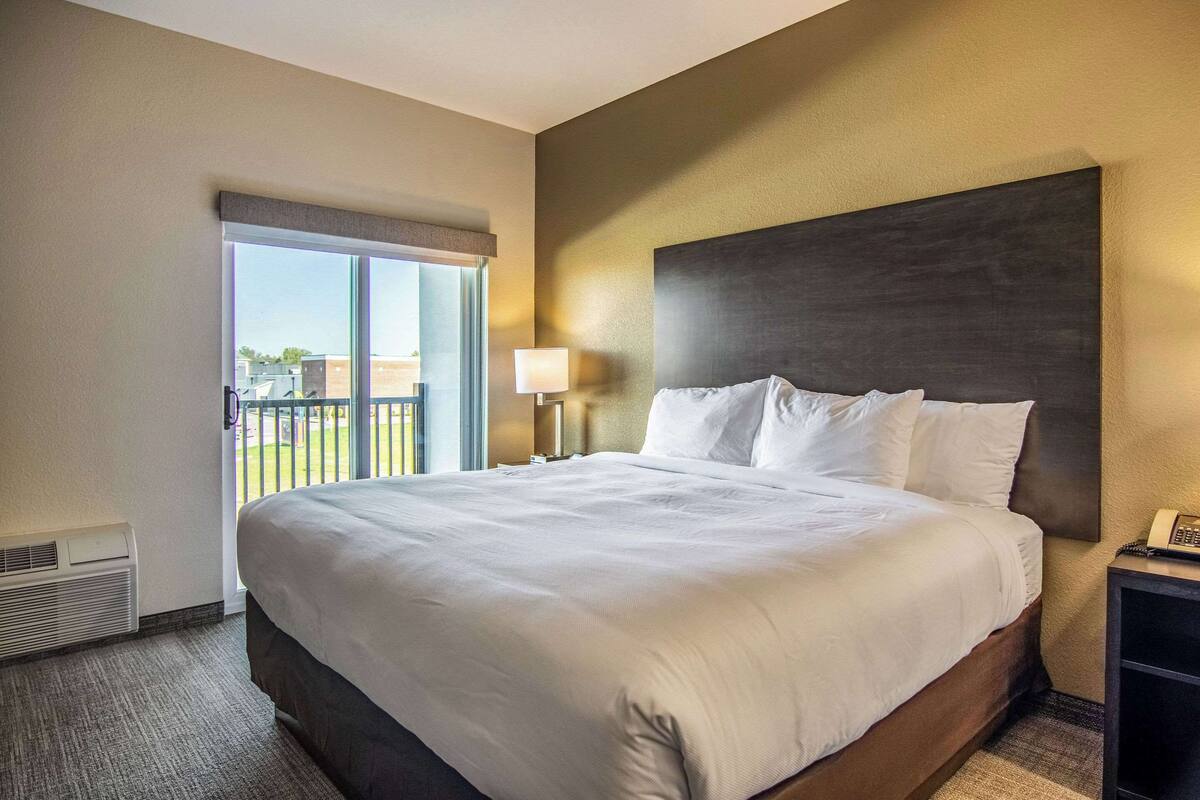 A hotel room with a large bed, a nightstand with a phone, a window with blinds, and a view of the outside.