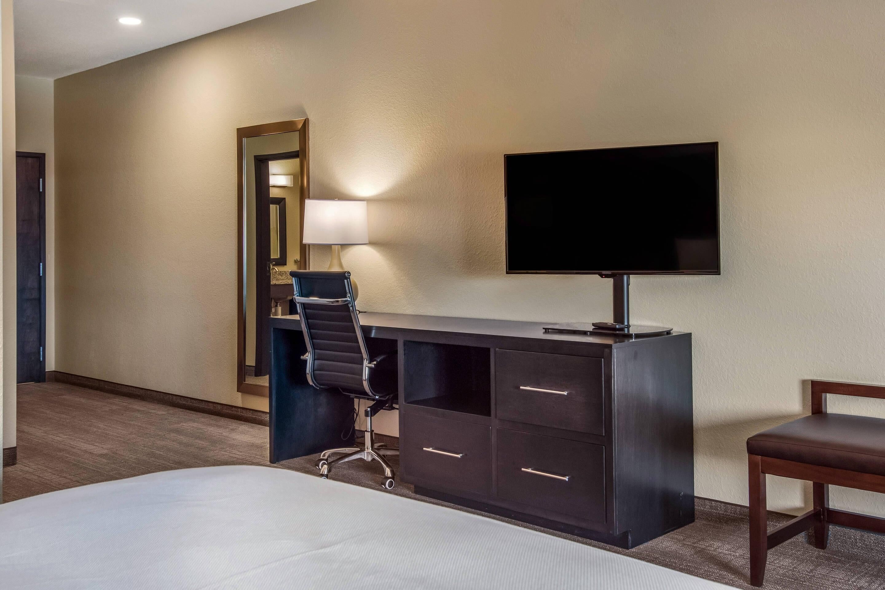 Una habitación de hotel con cama, escritorio, silla, televisor y espejo.