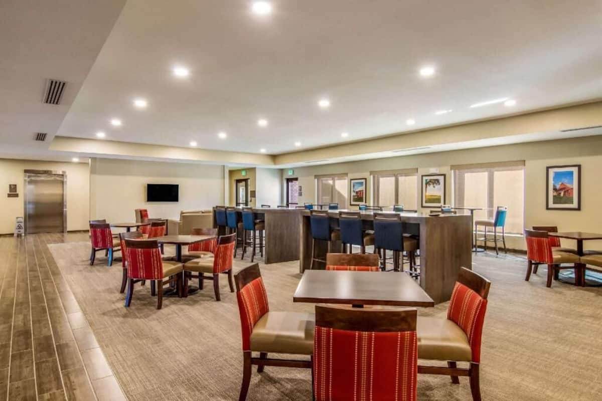 A hotel dining area with multiple tables and chairs, a central bar, and a kitchen in the background.