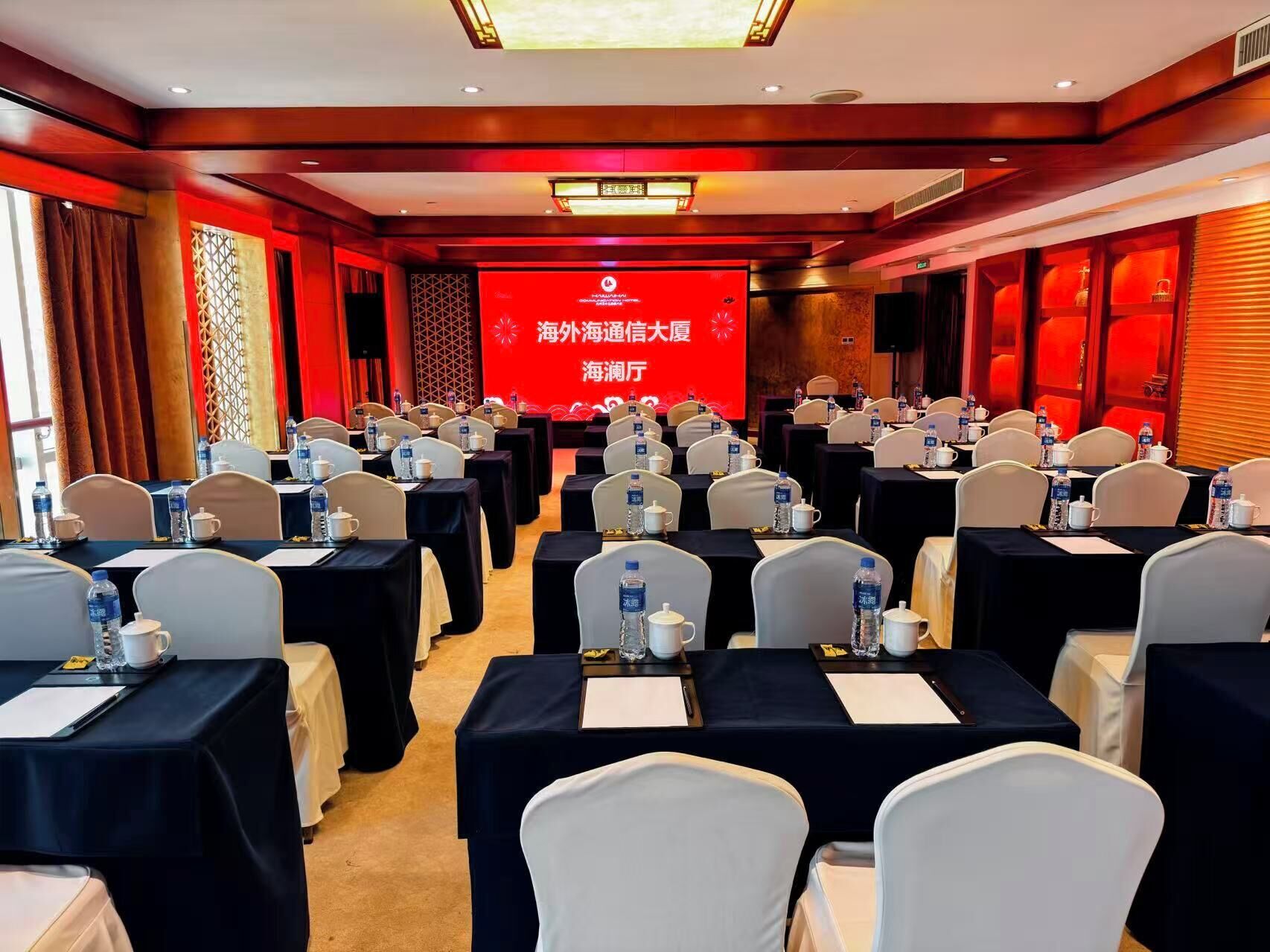 Un salón de conferencias con filas de mesas, sillas y una pantalla grande al frente.
