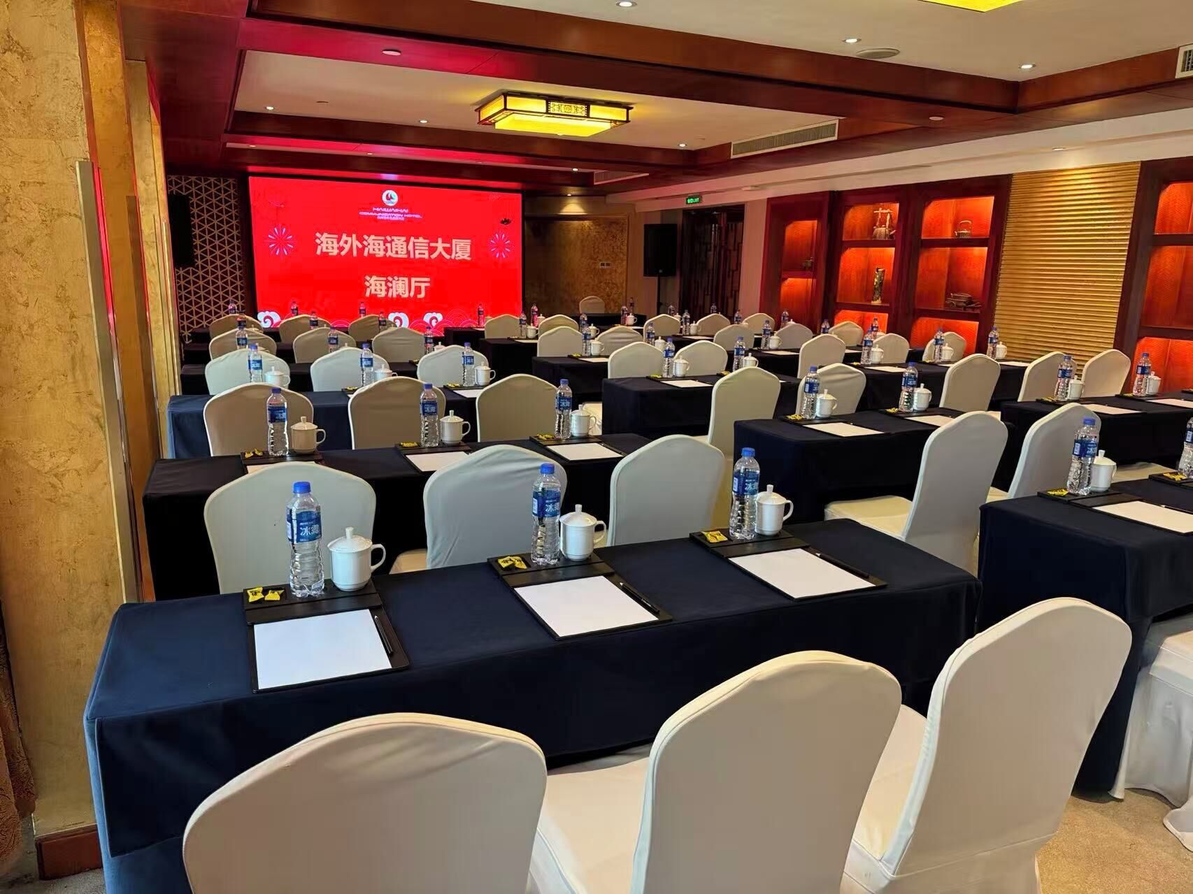 Un salón de conferencias con filas de mesas, sillas y una pantalla grande mostrando texto.