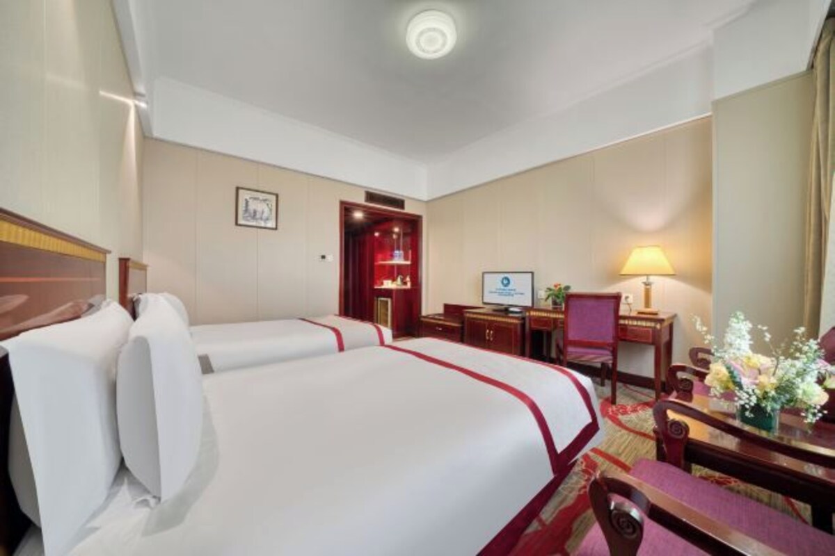 Habitación de hotel con dos camas, un escritorio con computadora, una silla, una lámpara de escritorio y una pequeña mesa con flores.