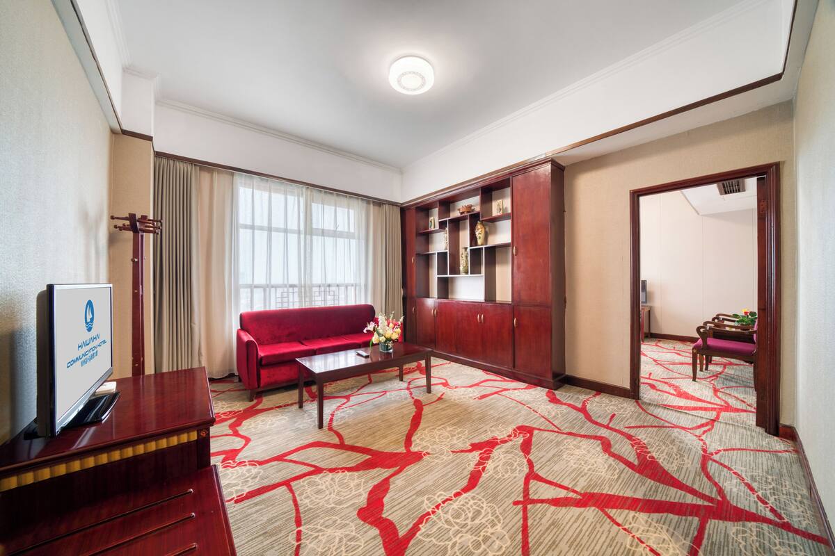 A hotel room with a red sofa, a wooden cabinet, a flat-screen TV, and a patterned carpet.