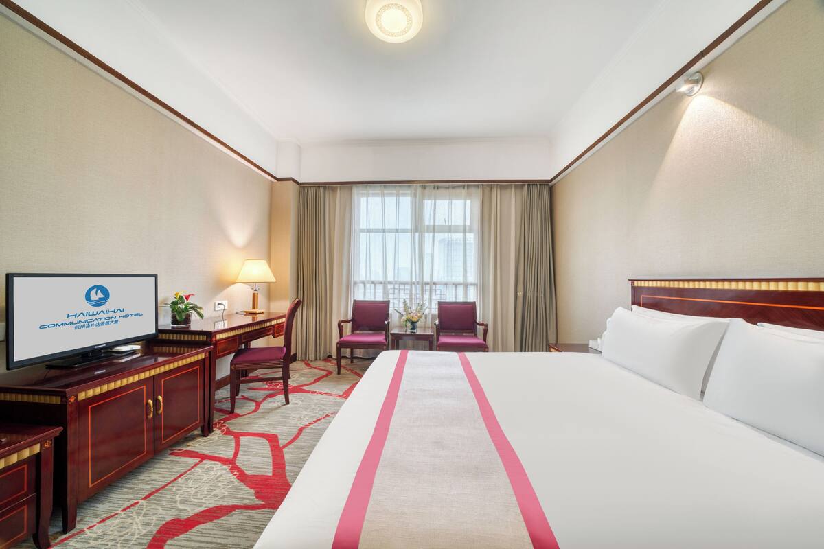 A hotel room with a large bed, a desk with a TV, two chairs, a lamp, and a window with curtains.