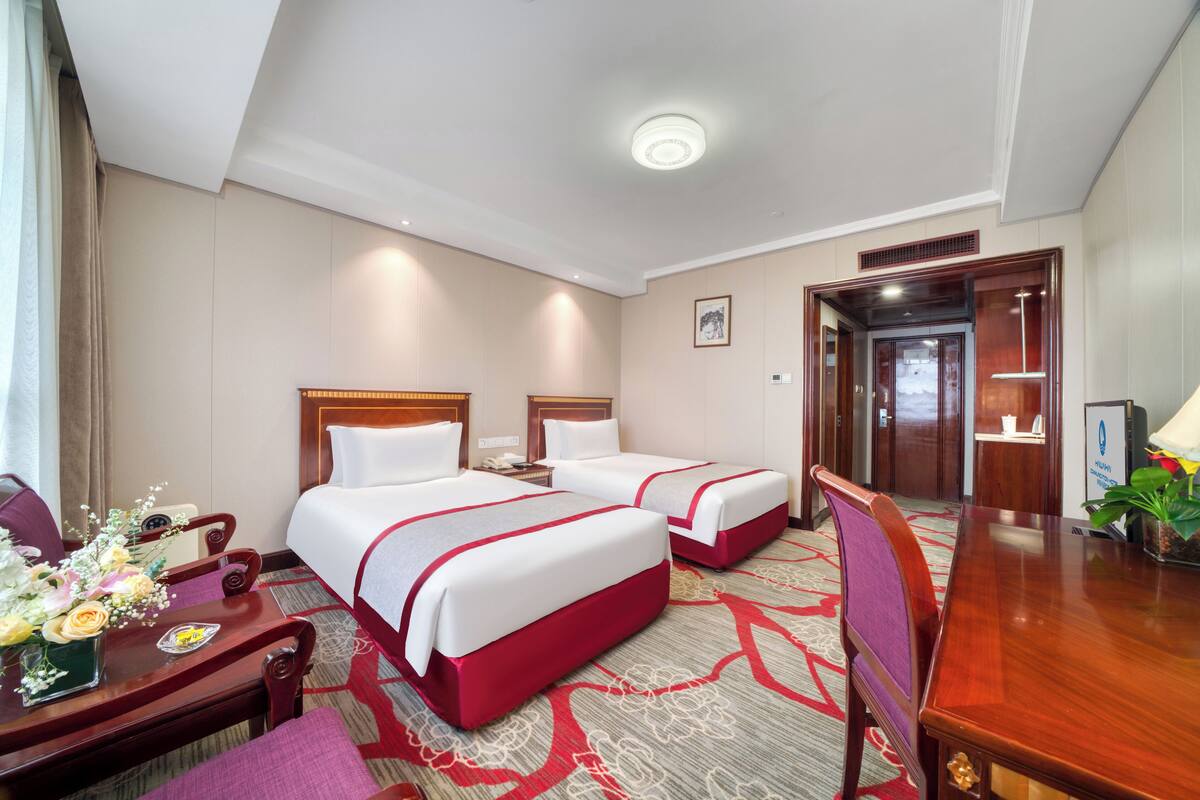 A hotel room with two beds, a desk, a chair, a table with a vase of flowers, and a door leading to a bathroom.