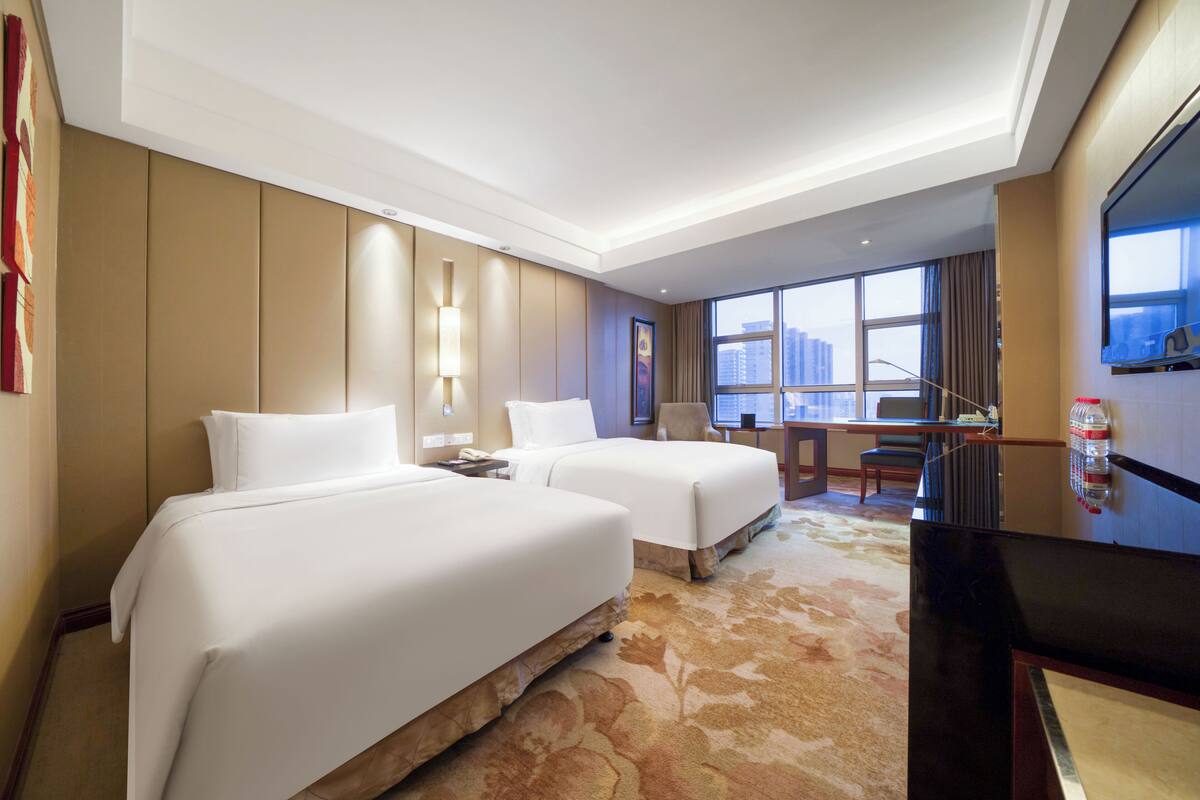 A hotel room with two beds, a desk, a chair, a TV, and a large window with a city view.