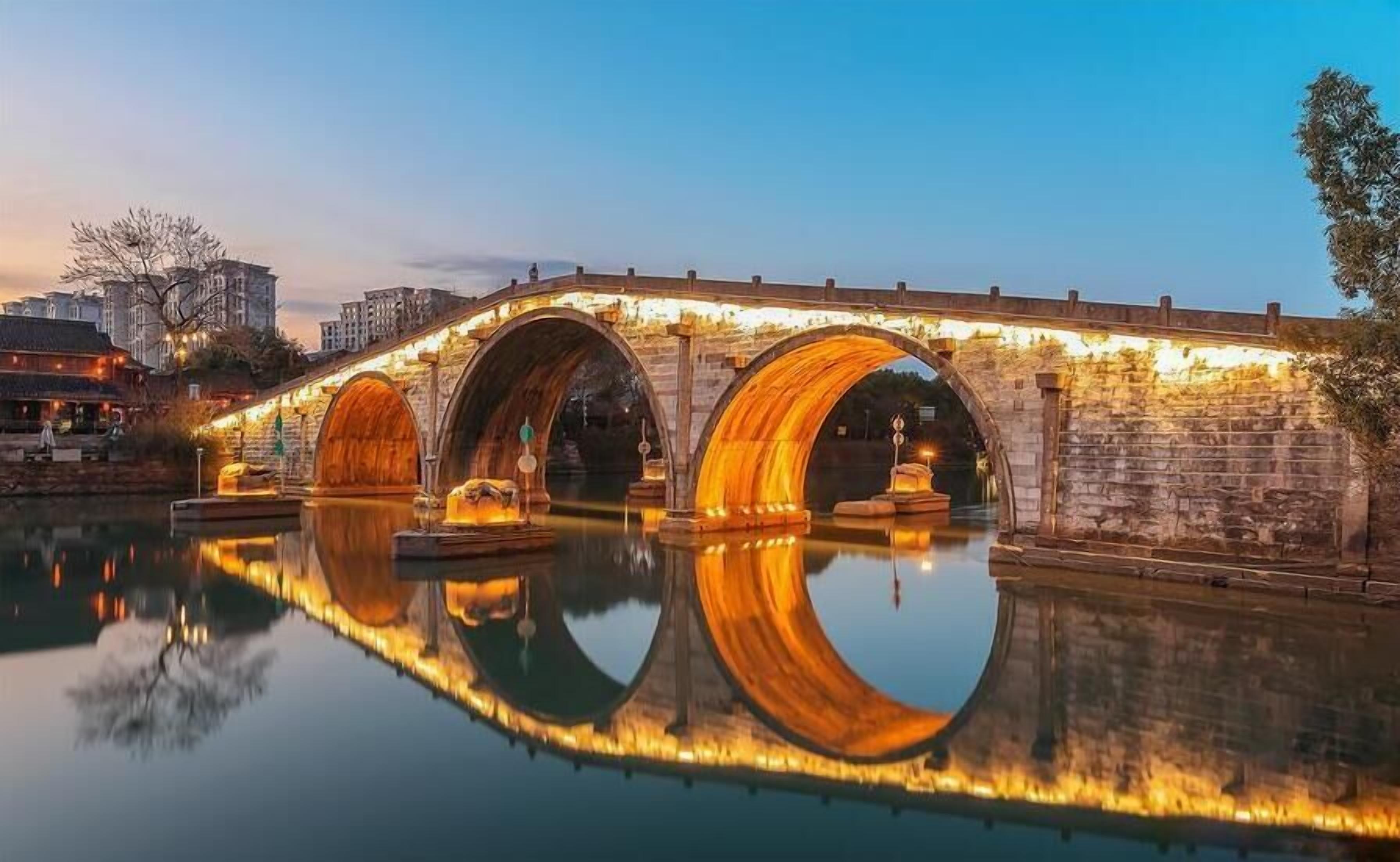 Un puente de piedra con tres arcos sobre un cuerpo de agua tranquilo, que refleja el puente y los edificios circundantes.