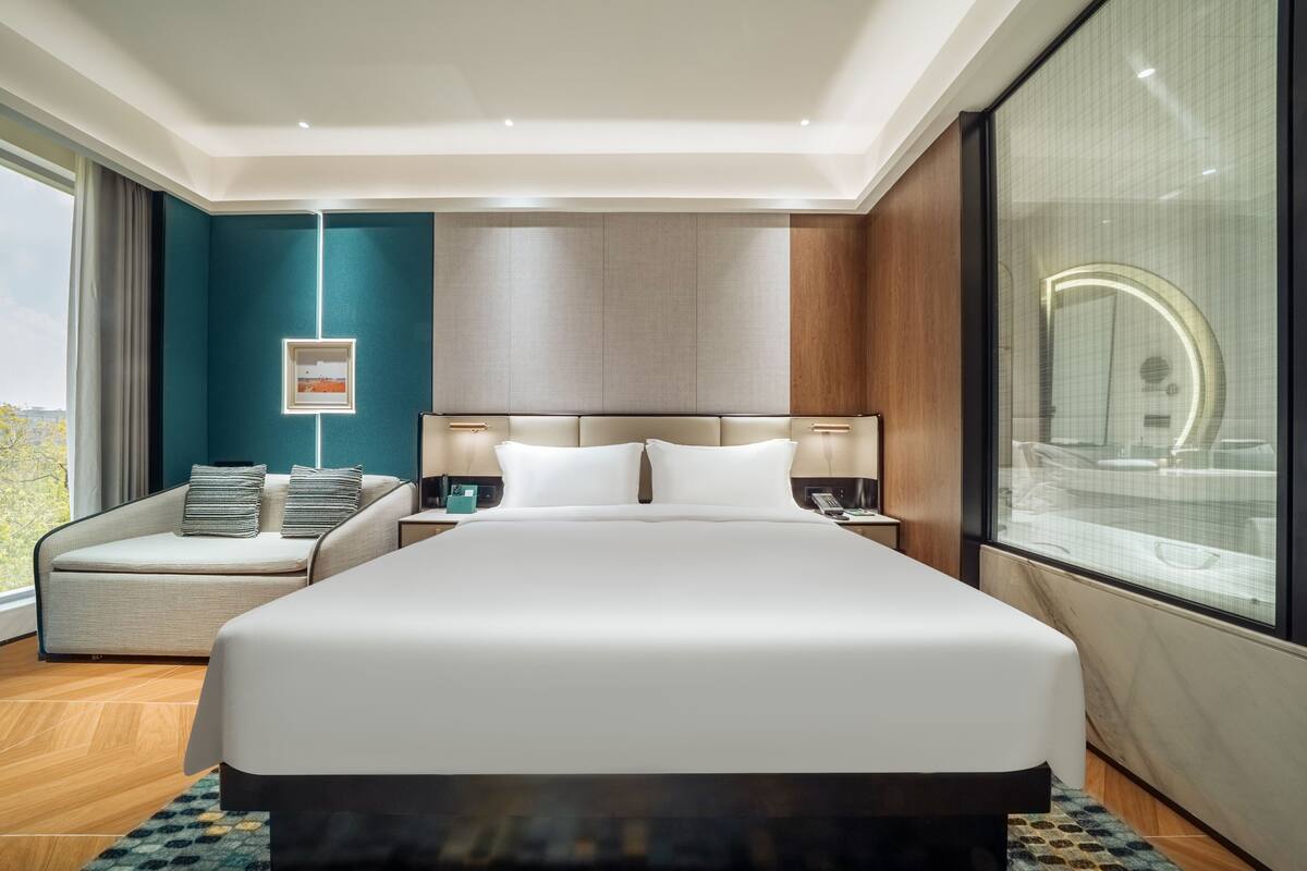 A modern hotel room with a large bed, a sofa, a nightstand, and a window with a view.