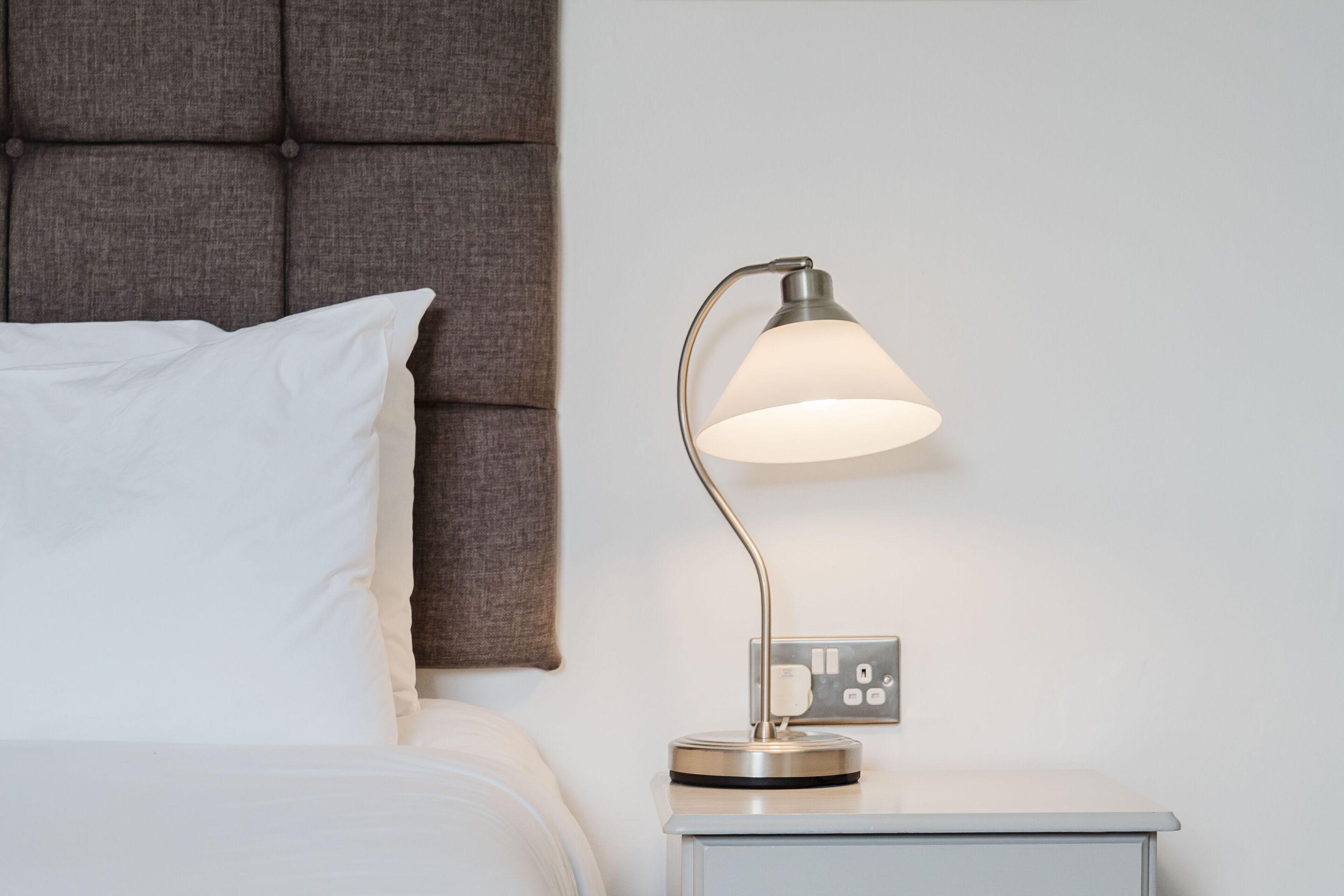 A neatly made bed with white linens and a grey headboard, a bedside table with a modern lamp, and a power outlet on the wall.