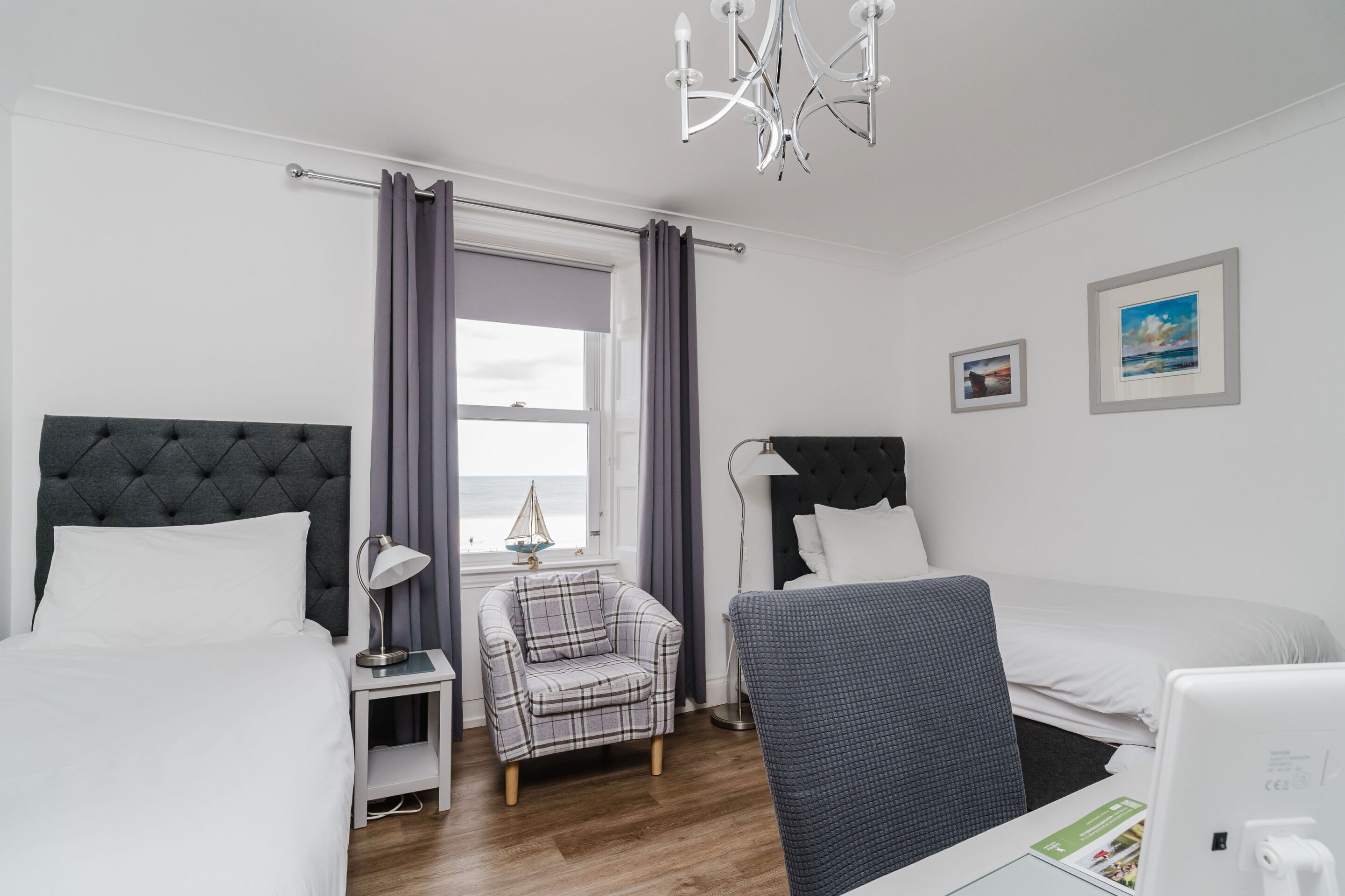 A hotel room with two beds, a chair, a desk, a lamp, and a view of the sea.