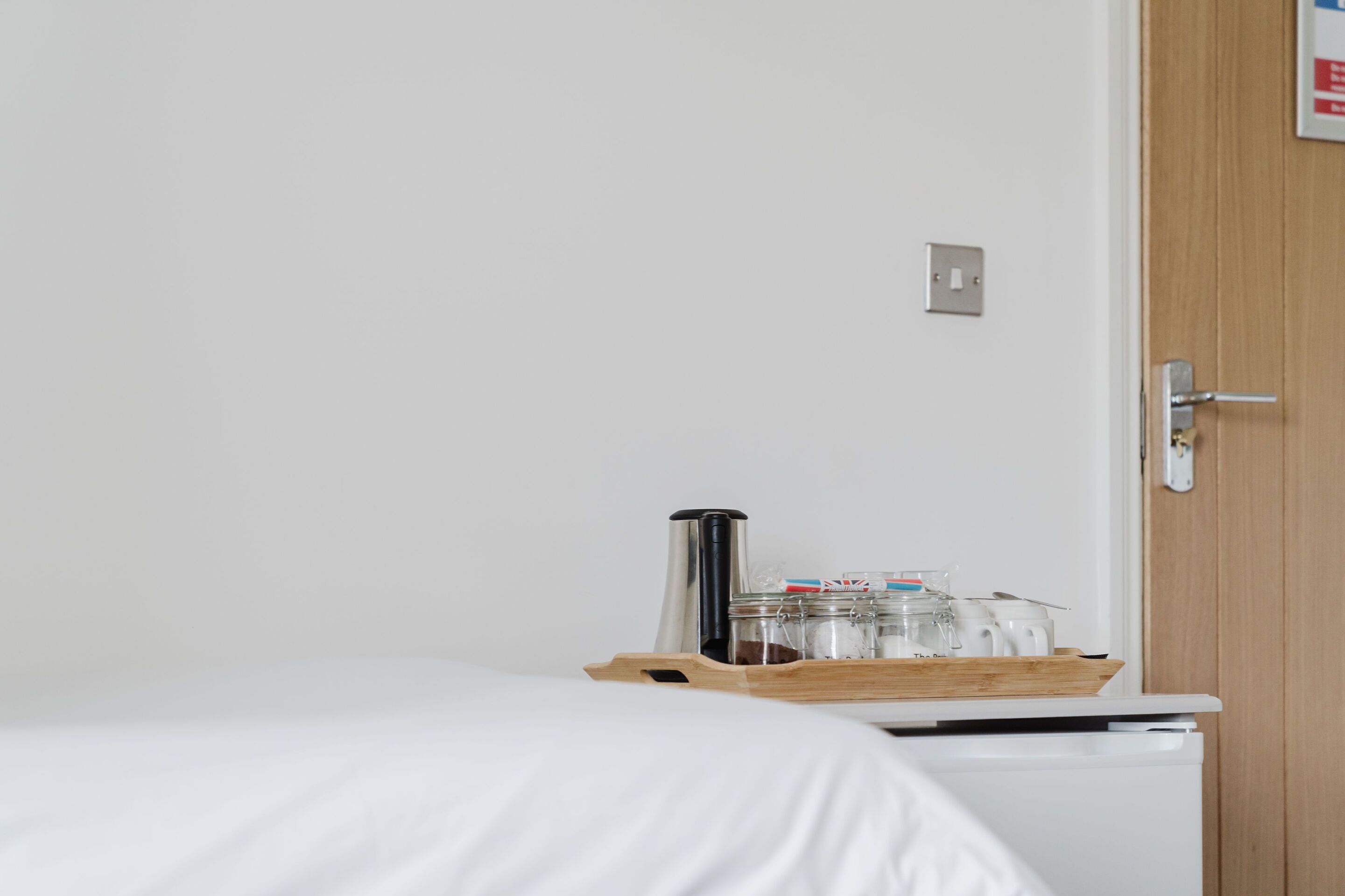 A hotel room with a bed, a wooden tray with a coffee pot and jars, and a door with a handle.