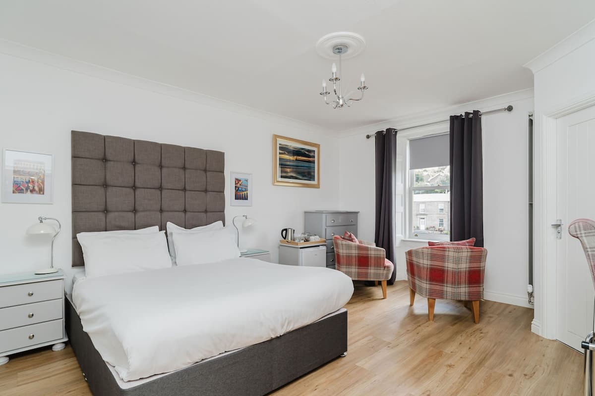 A bedroom with a large bed, a bedside table, a chair, a desk, and a window with curtains.