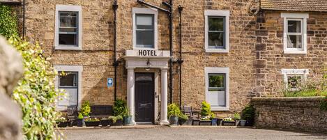 A stone building with a 'HOTEL' sign above the entrance, multiple windows, and potted plants in front.