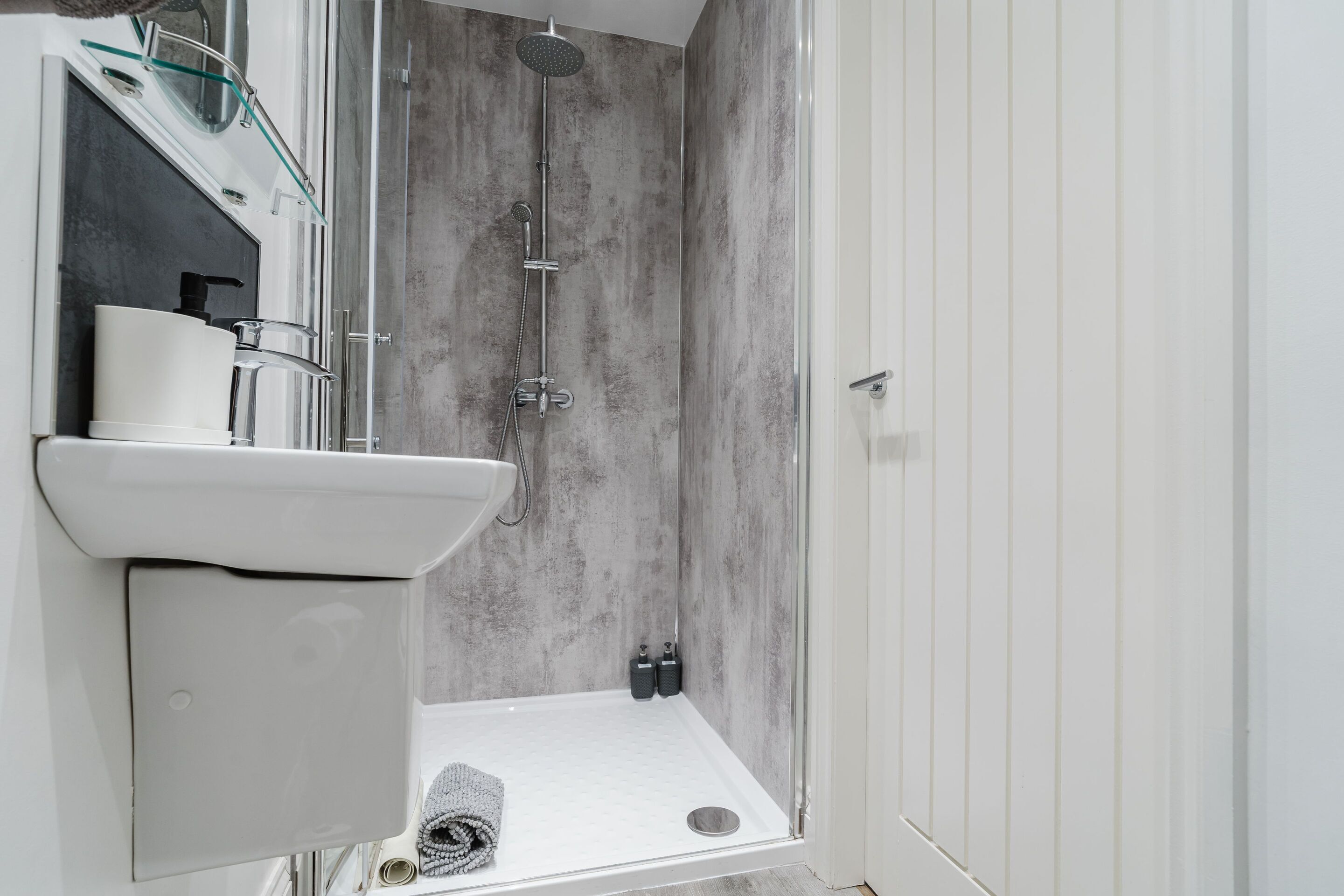 A modern bathroom with a glass shower enclosure, a white sink, and a towel on the floor.