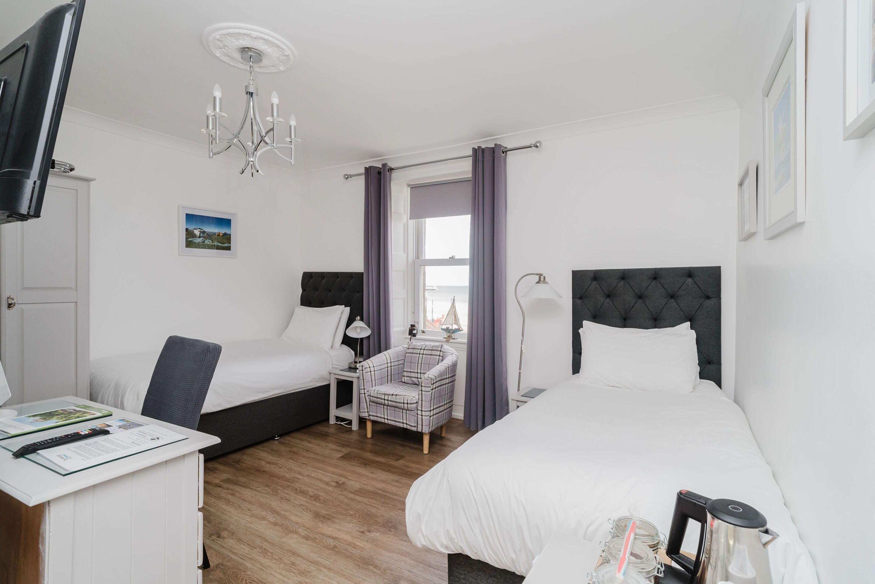A hotel room with two beds, a desk, a chair, a TV, a chandelier, and a window with curtains.