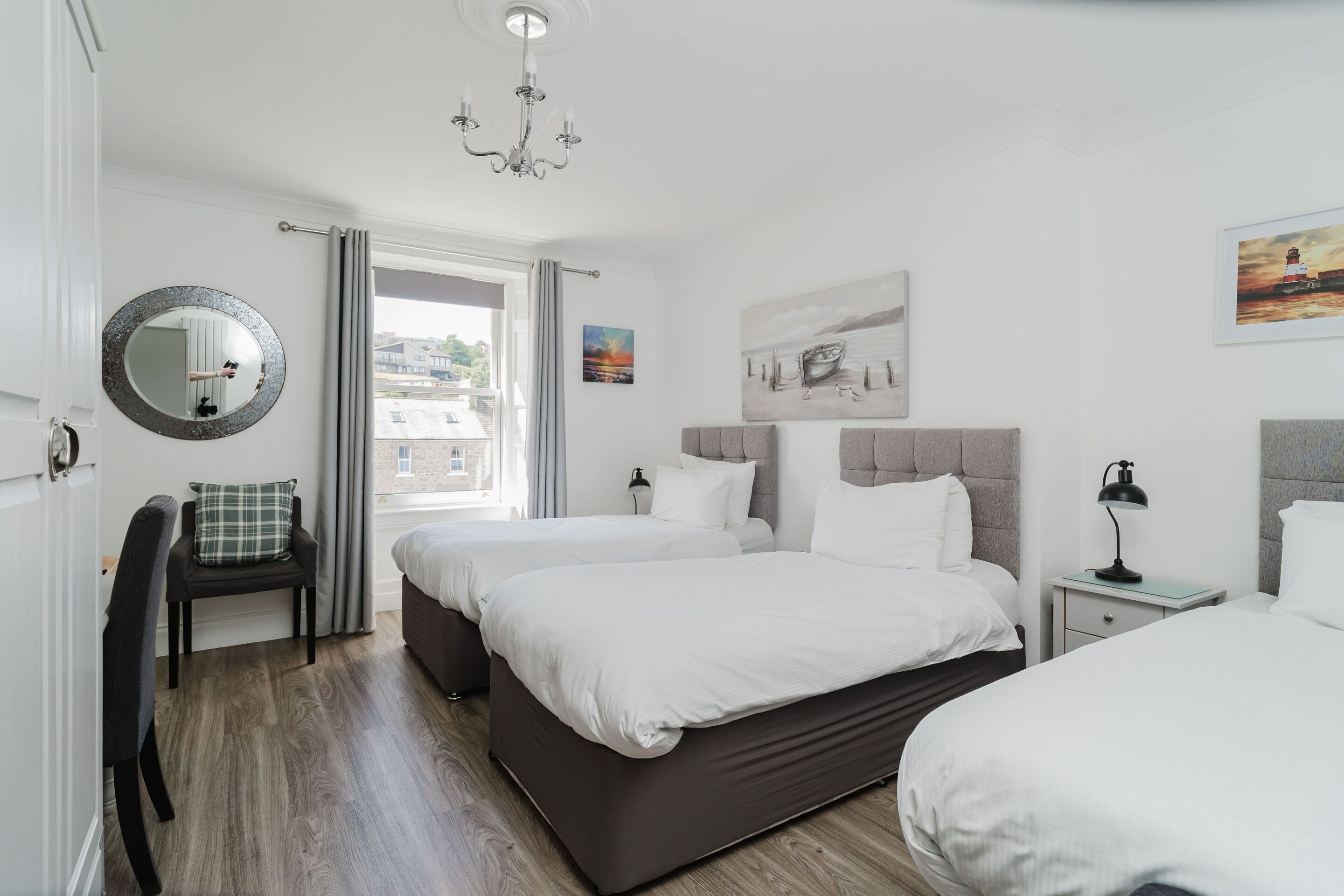 A modern hotel room with two beds, a round mirror, a chair, a nightstand, and artwork on the walls.