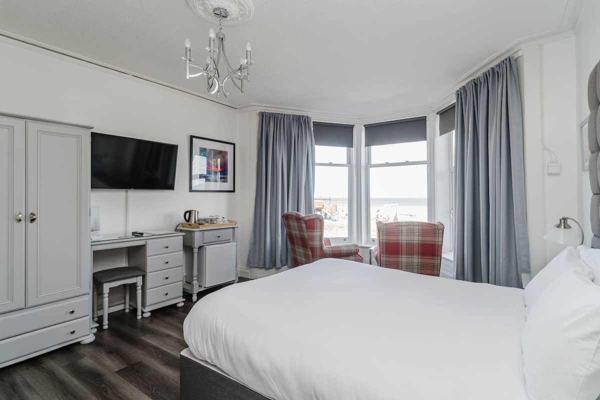 A hotel room with a large bed, a desk with a chair, a television, and a view of the sea through the windows.