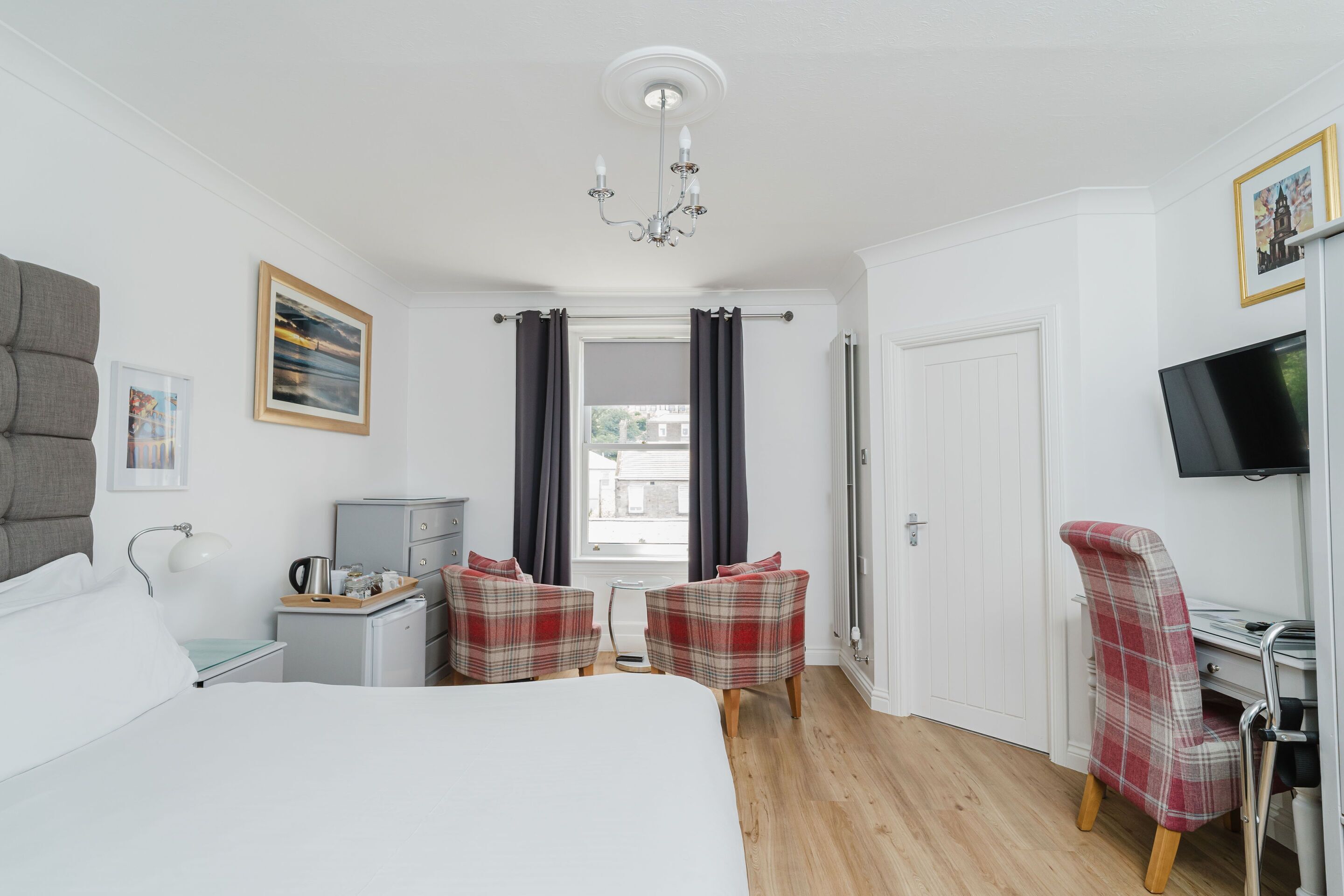 A hotel room with a bed, two plaid chairs, a desk with a chair, a television, a small table with a kettle, and a picture frame on the wall.
