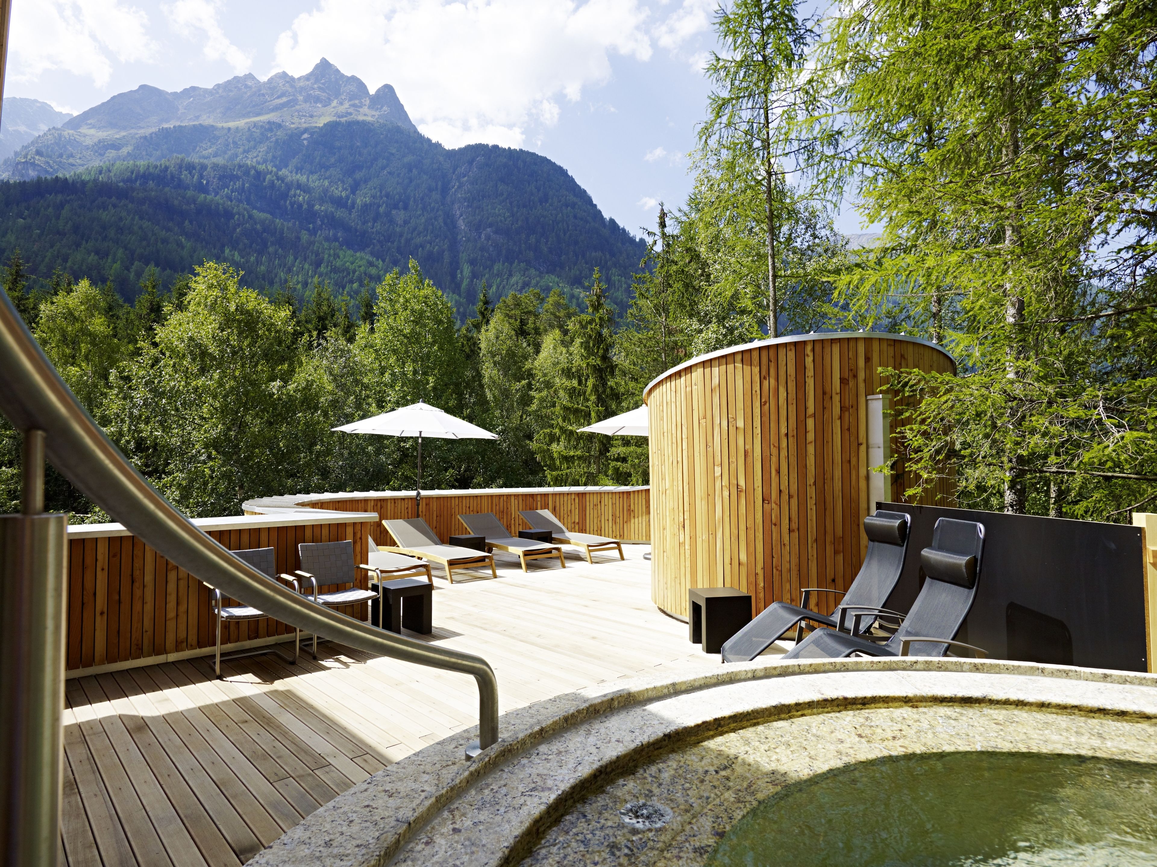 Terraza de madera con sillones de descanso y jacuzzi, con vistas a una cadena montañosa boscosa.