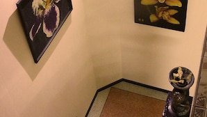 Μια σκάλα με ένα στηρίγμα για το χέρι, μια κορνιζαρισμένη εικόνα με λουλούδια στον τοίχο και ένα μικρό φυτό σε γλάστρα στο διάδρομο ανάμεσα στα σκαλοπάτια.