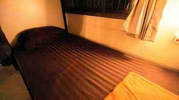 A single bed with a dark-colored mattress and a pillow, a small orange cushion, and a window with curtains.