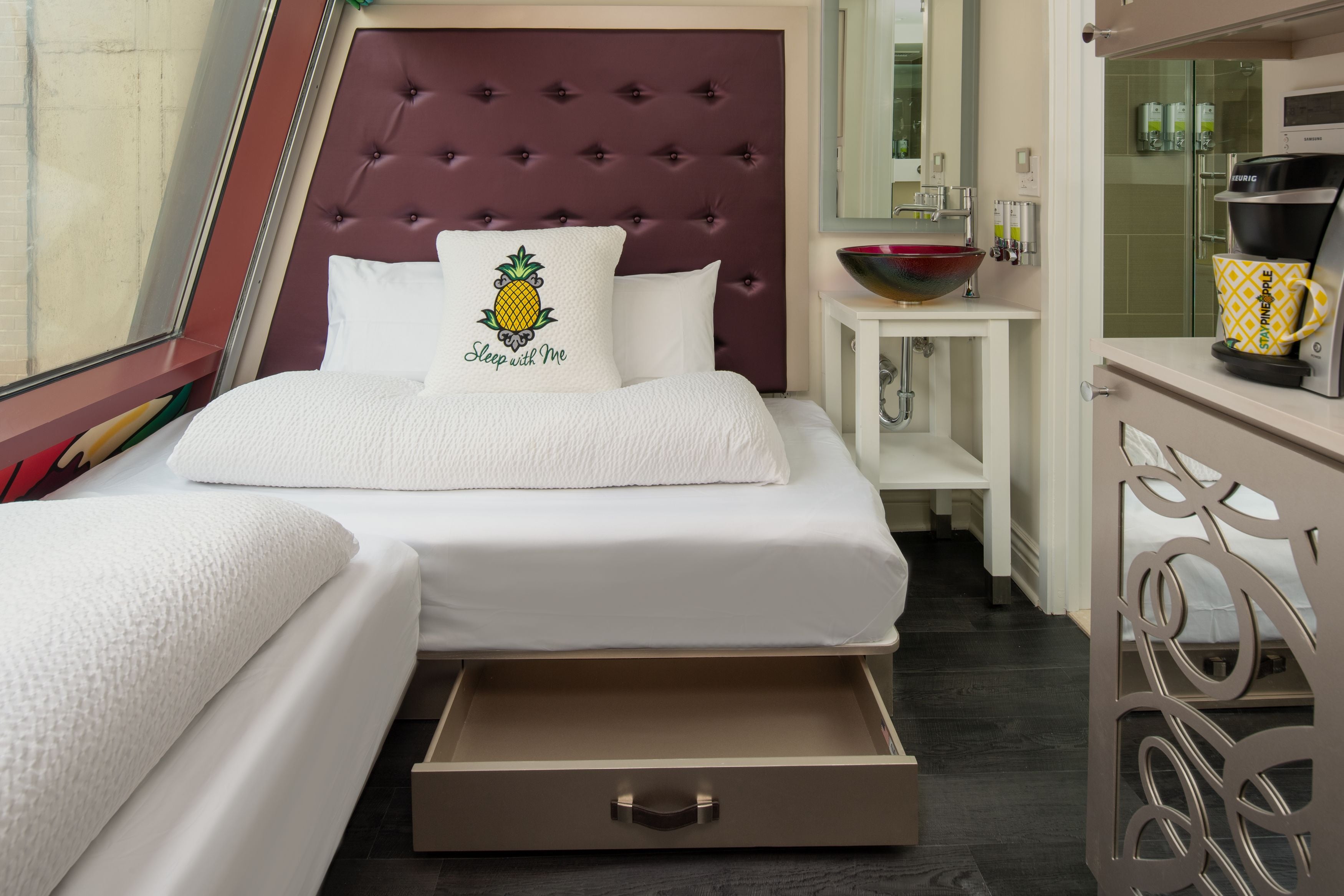 Una habitación de hotel con un cabecero capitonado, una cama con una colcha blanca y un cojín con diseño de piña, una mesita de noche con un cuenco y una cafetera, y un cajón debajo de la cama.