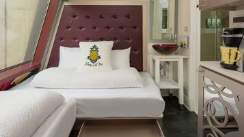 Una habitación de hotel con un cabecero capitonado, una cama con una colcha blanca y un cojín con diseño de piña, una mesita de noche con un cuenco y una cafetera, y un cajón debajo de la cama.