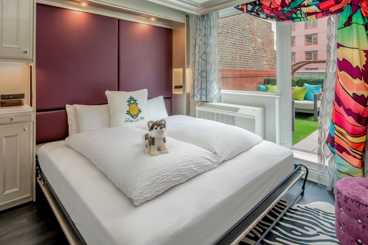 A hotel room with a bed, a plush toy, a window with a view of a building, and a balcony with a colorful curtain.