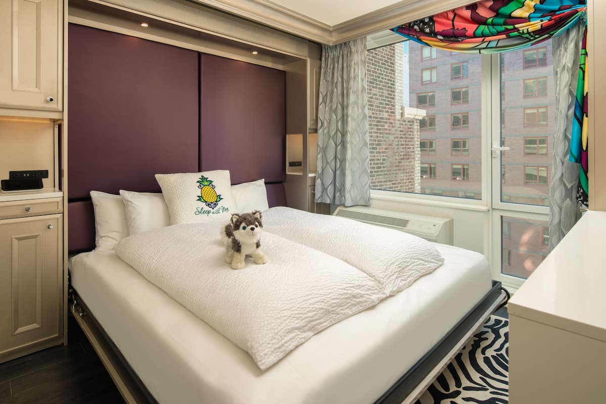 A neatly made bed with a plush pillow and a small stuffed animal, a window with patterned curtains, and a view of a building outside.